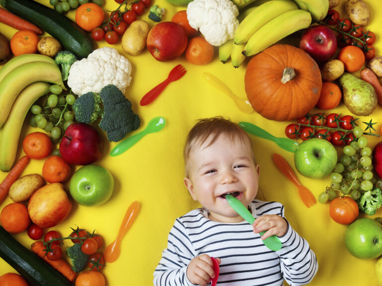 Bebé feliz rodeado de frutas y verduras variadas, alimentos fundamentales en la dieta infantil de 1 a 3 años