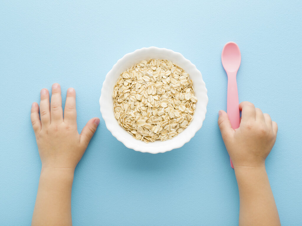 Manos de bebé junto a un bol de copos de avena y cuchara, cereales en la alimentación infantil