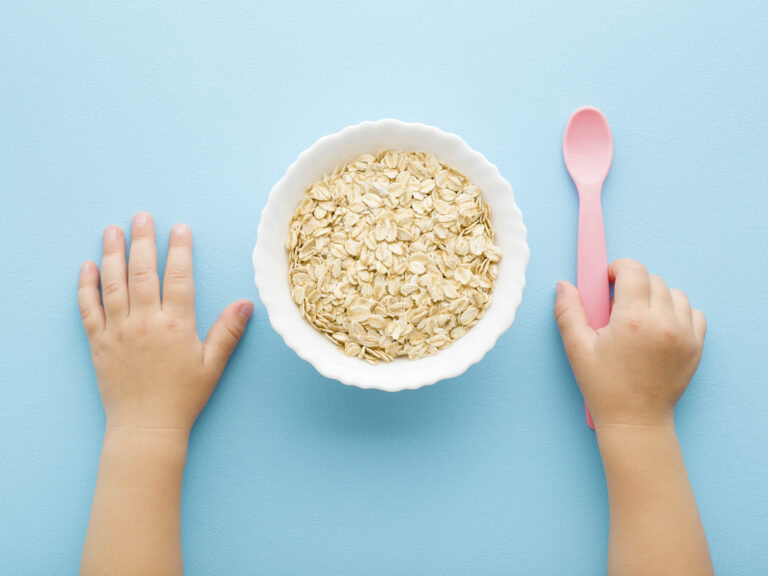 Manos de bebé junto a un bol de copos de avena y cuchara, cereales en la alimentación infantil