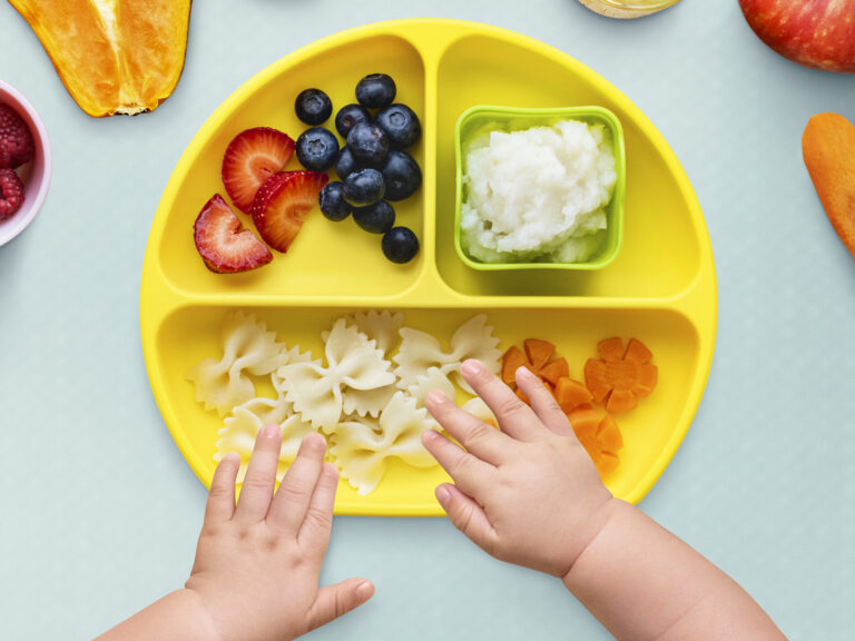 Manos de bebé sobre plato infantil con pasta, zanahoria, fresas y arándanos, distribución equilibrada de alimentos para niños de 1 a 3 años