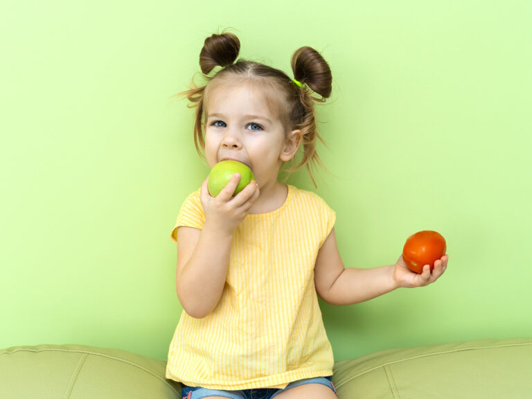 Niña comiendo una manzana verde y sosteniendo una naranja, fomentando el consumo de fruta en niños