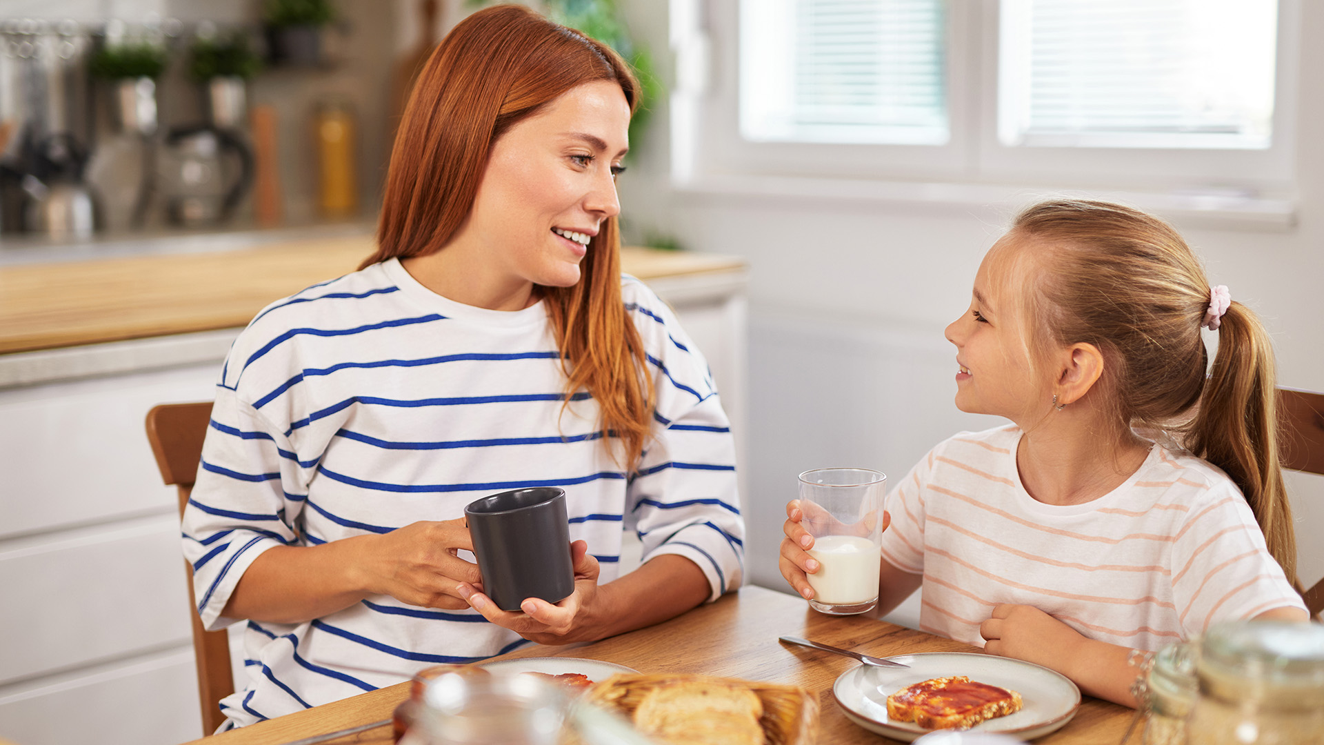 Madre e hija desayunando juntas en cocina con leche y pan tostado