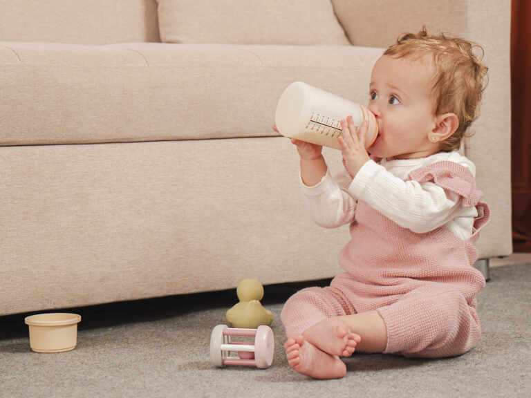 Bebé vestida de rosa sentada en el suelo rodeada de juguetes, bebiendo de un biberón de alimento lácteo, en el contexto de la dieta infantil de 6 a 24 meses