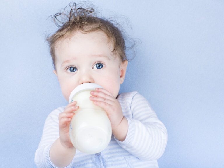Bebé de ojos azules sosteniendo y bebiendo de un gran biberón de fórmula láctea de continuación, sobre fondo azul claro