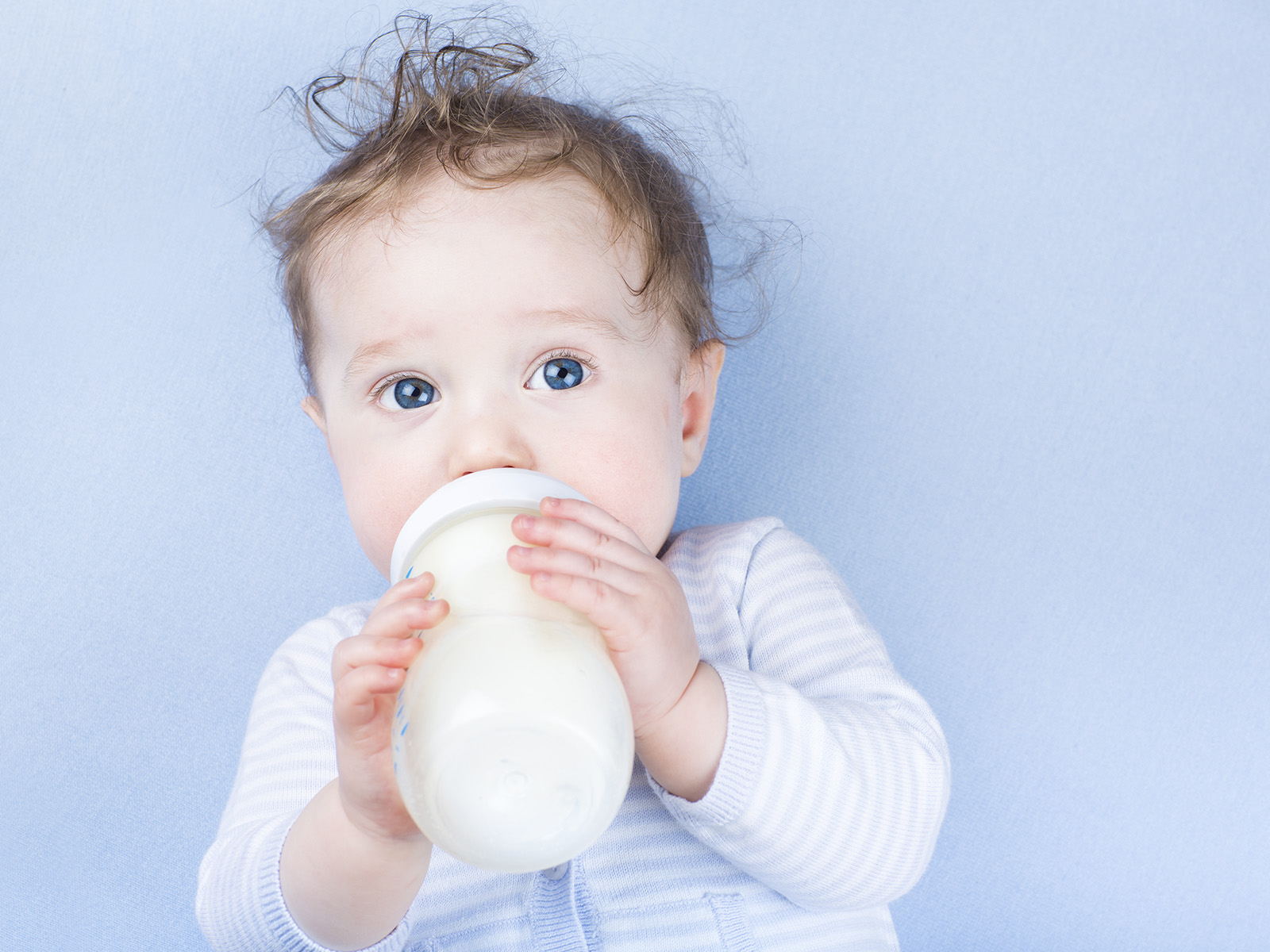 Bebé de ojos azules sosteniendo y bebiendo de un gran biberón de fórmula láctea de continuación, sobre fondo azul claro
