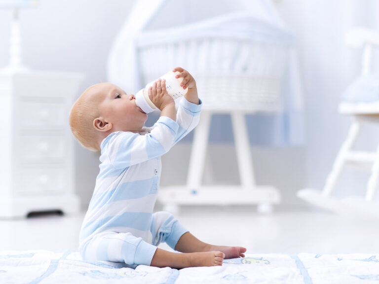 Bebé tomando un biberón de leche de inicio sentado sobre una manta blanca, con una cuna de madera al fondo en una habitación clara