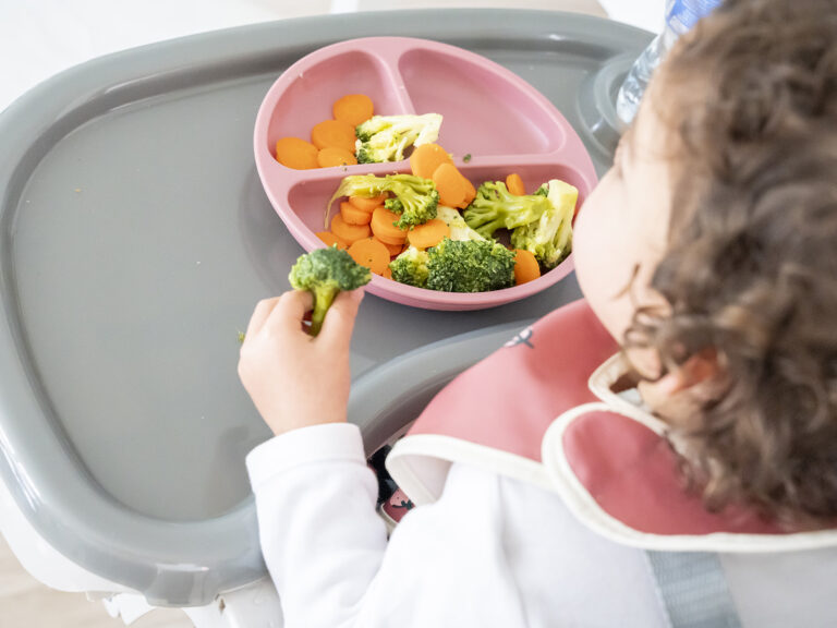 Bebé sentado en su trona comiendo brócoli y zanahorias en un plato rosa con divisiones, promoviendo la alimentación saludable con legumbres y verduras desde la infancia