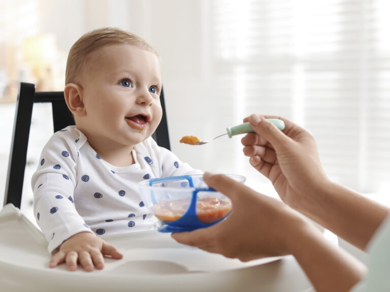 Bebé de ojos azules con la boca abierta siendo alimentado con una cuchara de papilla de frutas o verduras, ilustrando la introducción de papillas según la edad del bebé