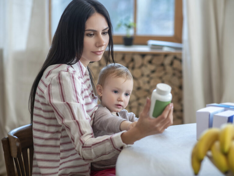 Madre alimentando a bebé pequeño con vaso de bebida nutritiva verde en cocina luminosa
