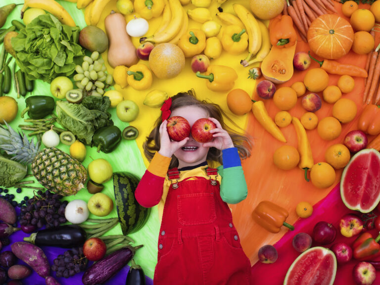 Niño jugando rodeado de frutas y verduras coloridas de arco iris, sosteniendo manzanas en los ojos